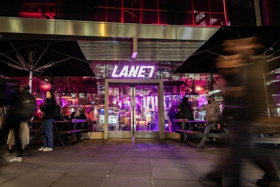 Exterior view of Lane7 bowling alley with a patio at night, with purple and red lights visible inside.
