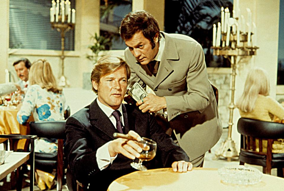 Actors Roger Moore and Tony Curtis from "The Persuaders!" in a restaurant scene, with Curtis holding money and Moore holding a cigar and glass.