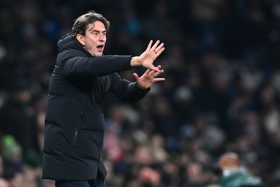 A man wearing a black puffer jacket and a red nose is gesturing with both hands, mouth open, against a blurred stadium background.