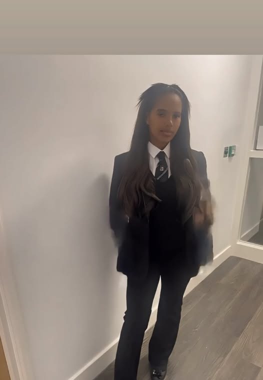 A young woman in a dark suit with a white shirt and tie stands in a hallway.