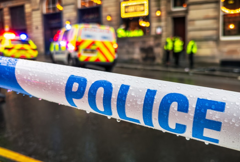 Close-up of "POLICE" barricade tape with raindrops, with police vehicles and officers blurred in the background.