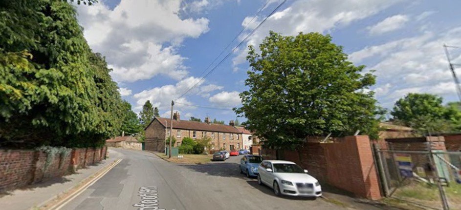 Street view of a road with parked cars, brick houses, and lush trees under a cloudy sky.