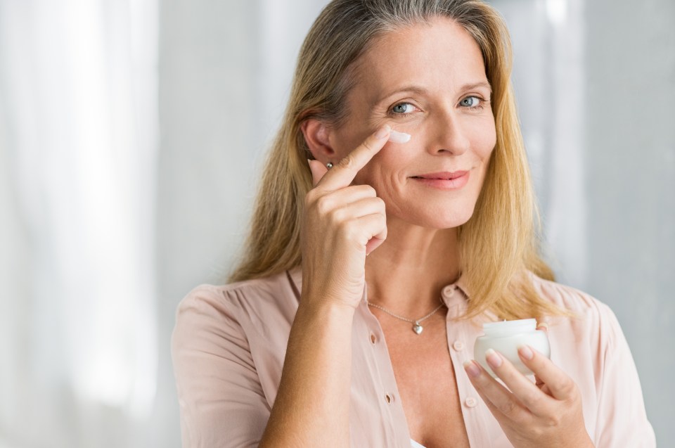 Smiling woman applying anti-aging lotion under her eyes while holding a jar of cream.
