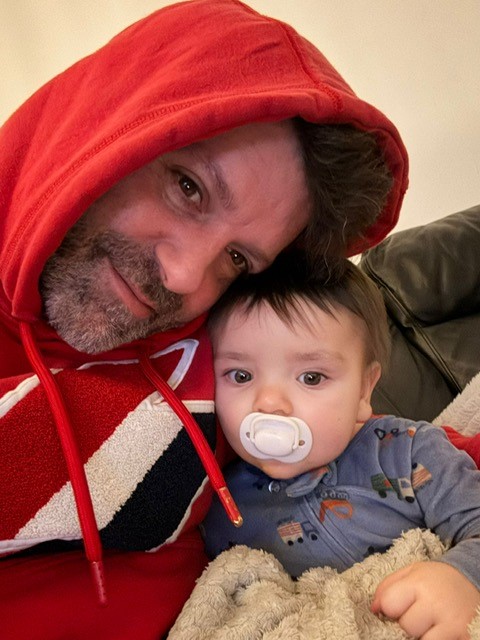 A man in a red hoodie leaning his head against a baby with a pacifier.