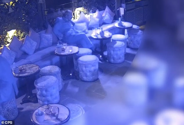 The reclining restaurant boss uses his right hand to reach into his trouser pocket and appears to pull out a small bottle filled with a substance