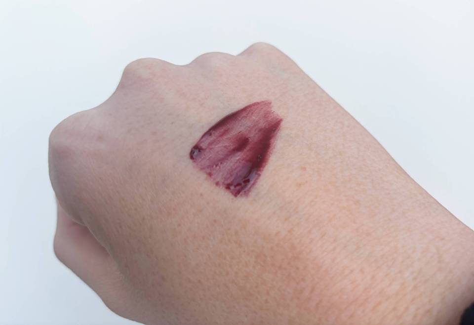 Swatch of Rimmel lip gloss in dark red with a glossy finish on fair skin.