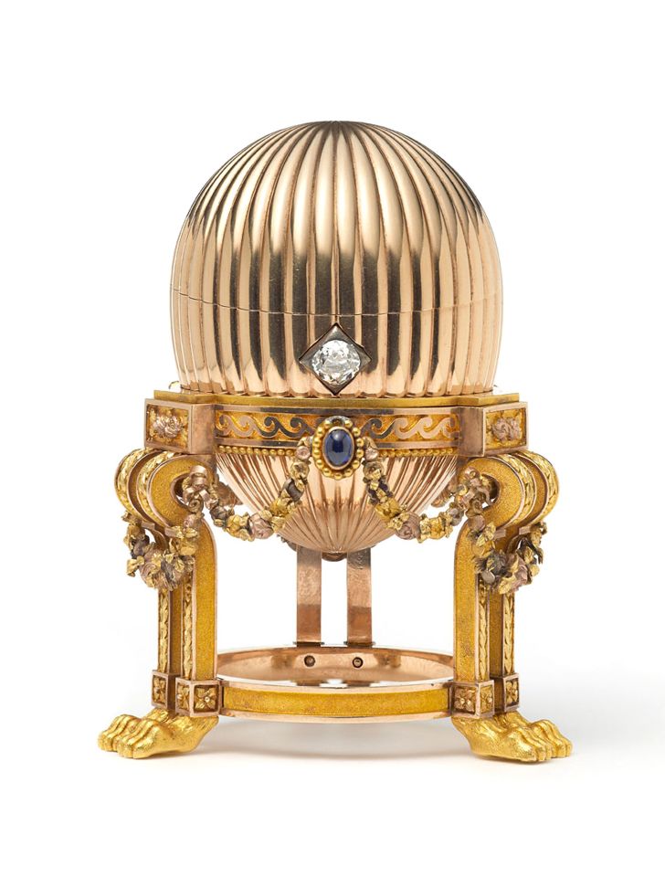A golden, ribbed Fabergé egg with a diamond and sapphire, on an ornate stand with lion's paw feet.