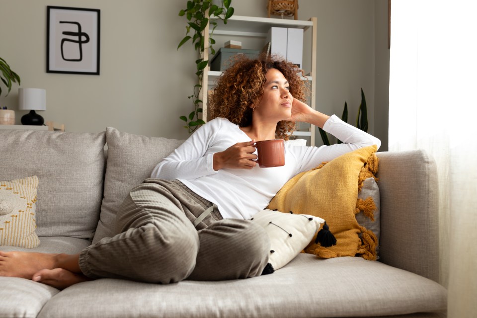 Pensive multiracial woman relaxing on a sofa, drinking morning coffee.