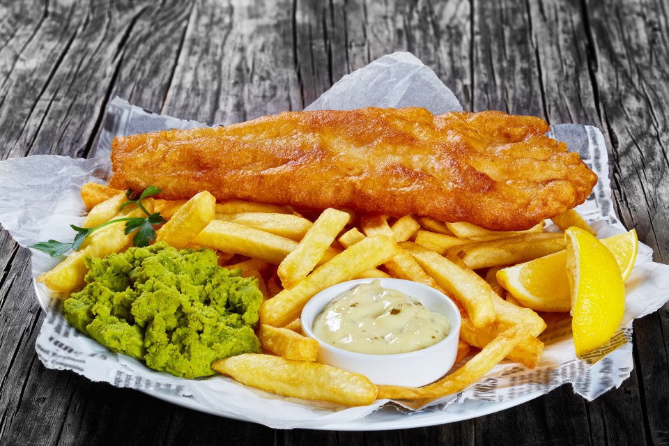Crispy fried cod, French fries, mushy peas, lemon slices, and tartar sauce served on newspaper atop a wooden table.