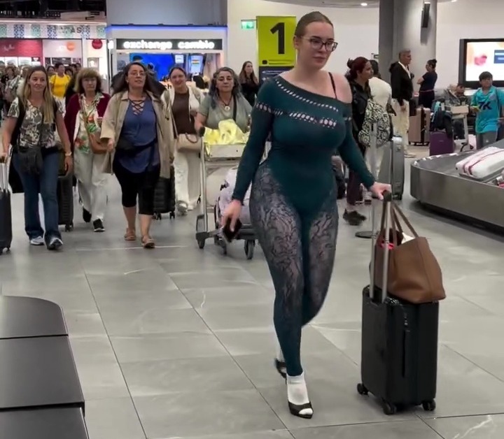 A woman in a revealing, form-fitting green and black outfit walking through an airport with a rolling suitcase.