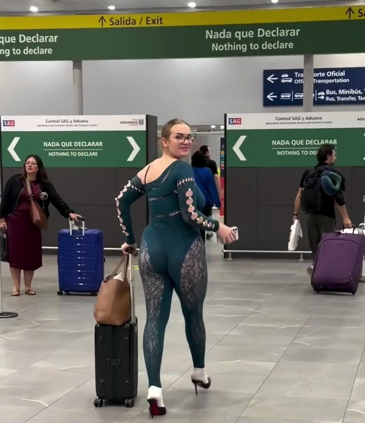 A traveler in a revealing dark green jumpsuit and high heels pulls a suitcase through an airport terminal.