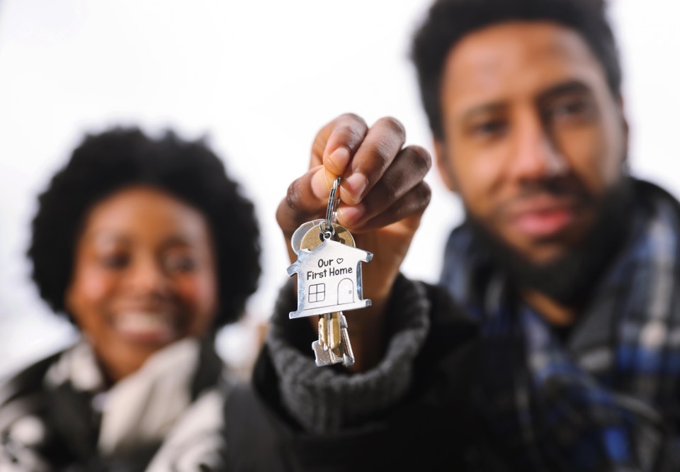A person holds up a house-shaped keychain that says "Our First Home", with another person smiling in the background.
