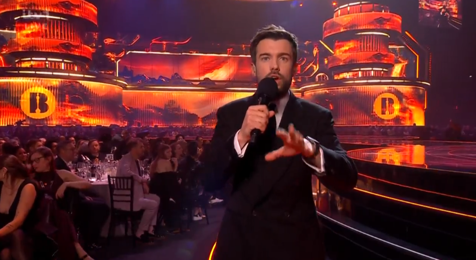 A man in a suit speaking into a microphone on stage at an awards ceremony, with a large "B" logo visible on the backdrop.