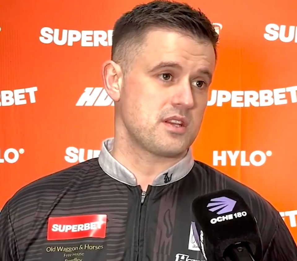 A man in a black and grey patterned shirt with sponsor logos from Superbet and Old Waggon & Horses speaking into a microphone with the Oche 180 logo.