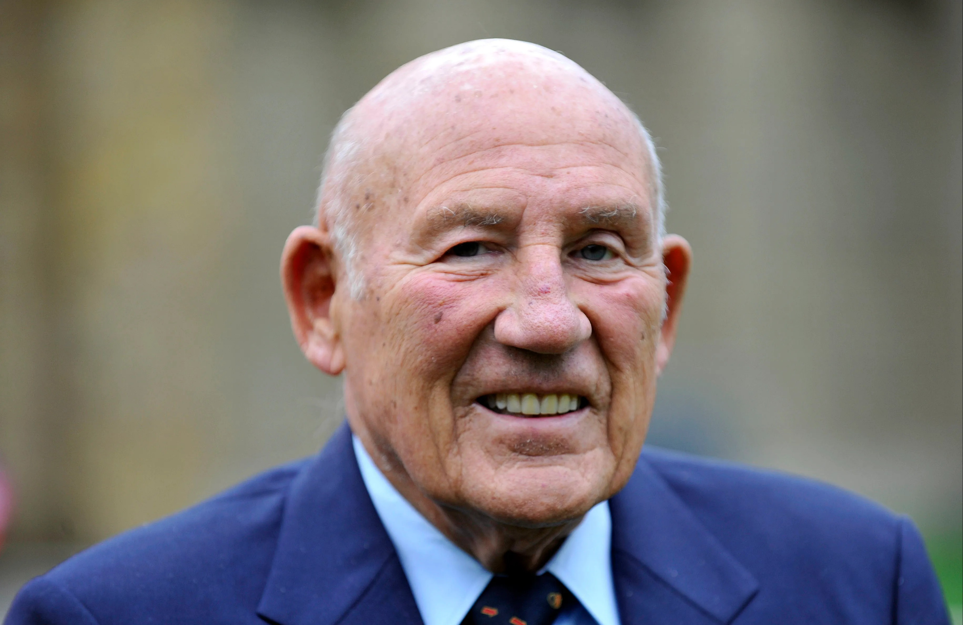 Former Formula One racing driver, Sir Stirling Moss, wearing a blue suit jacket and light blue collared shirt, smiling at the camera.