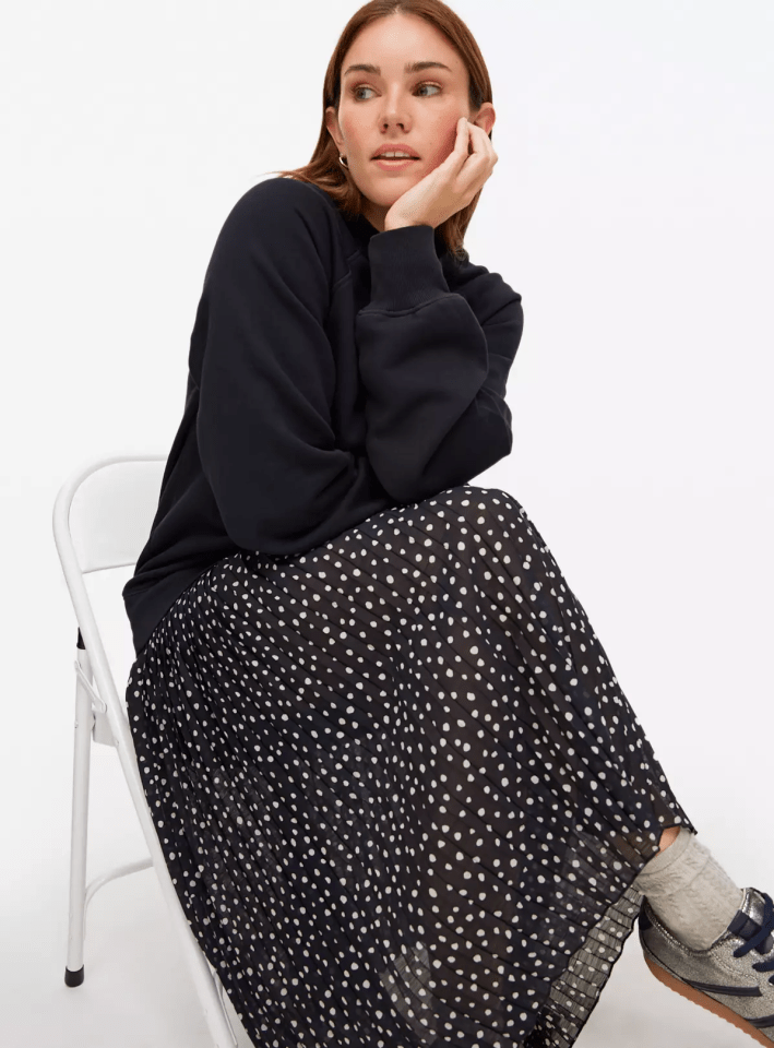 A woman wearing a black sweatshirt and black polka-dot skirt sits on a white chair.