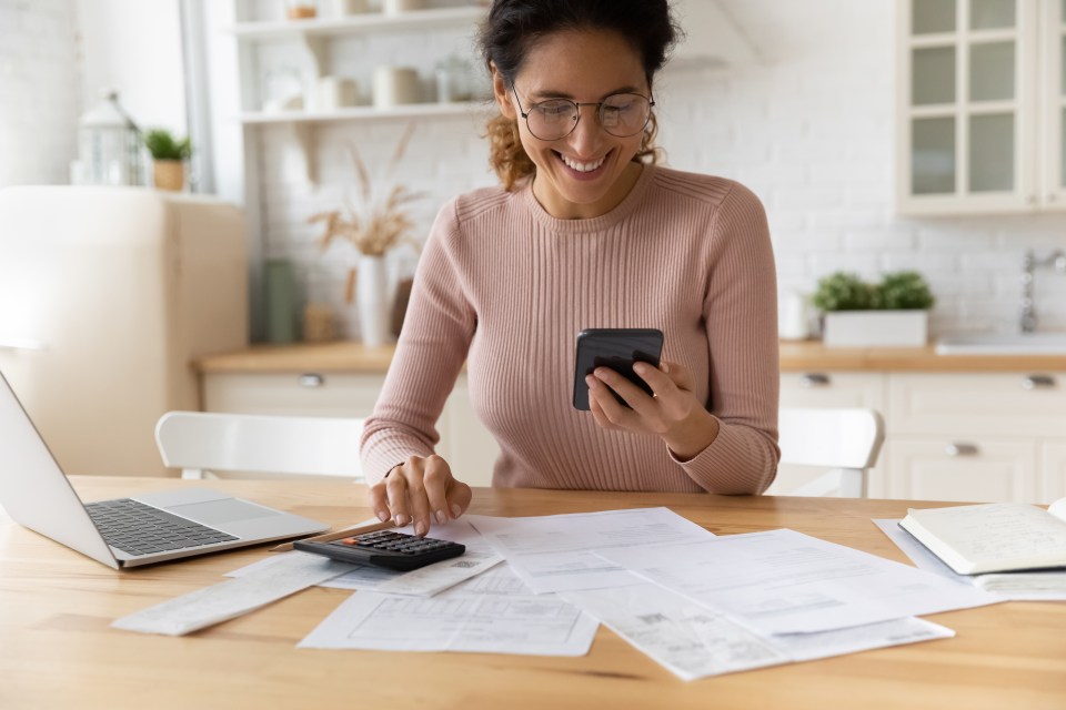 A smiling woman in glasses looks at her smartphone while calculating expenses at a table with a laptop and papers.