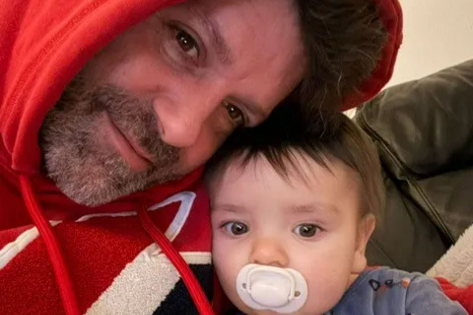 An image collage containing 1 images, Image 1 shows A man in a red hoodie leaning his head against a baby with a pacifier