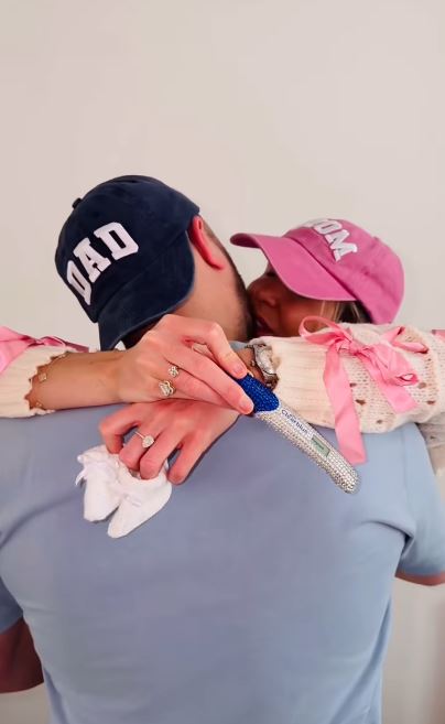A couple embracing, both wearing caps that say "MOM" and "DAD", holding a positive pregnancy test.