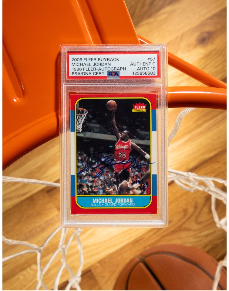 Michael Jordan's autographed 1986 Fleer-Autograph basketball card, authenticated by PSA, resting on a basketball hoop with a basketball visible below.