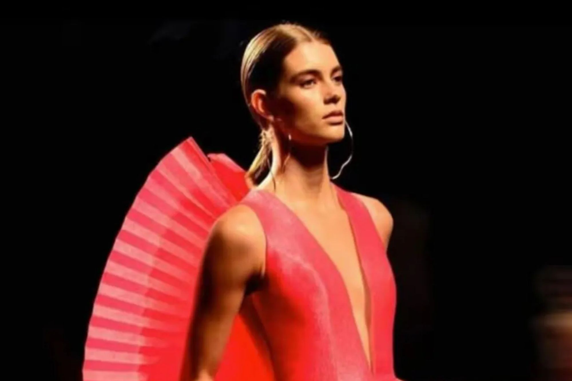 An image collage containing 1 images, Image 1 shows A fashion model with light brown hair, wearing a bright pink dress with a plunging V-neckline and a pleated fan-like detail on the shoulders, stands against a dark background