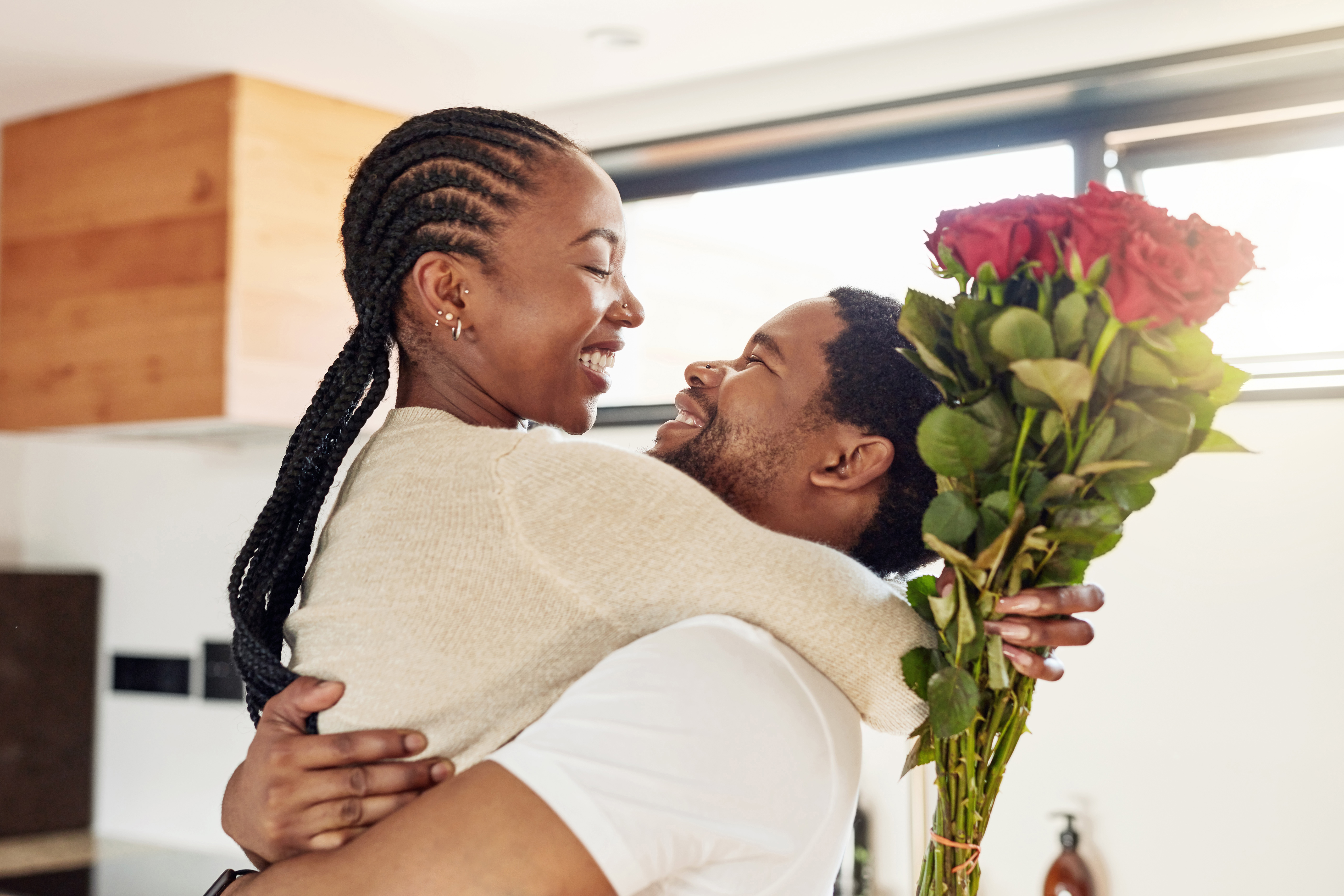 A young Black man lifts his smiling girlfriend who holds a bouquet of red roses.