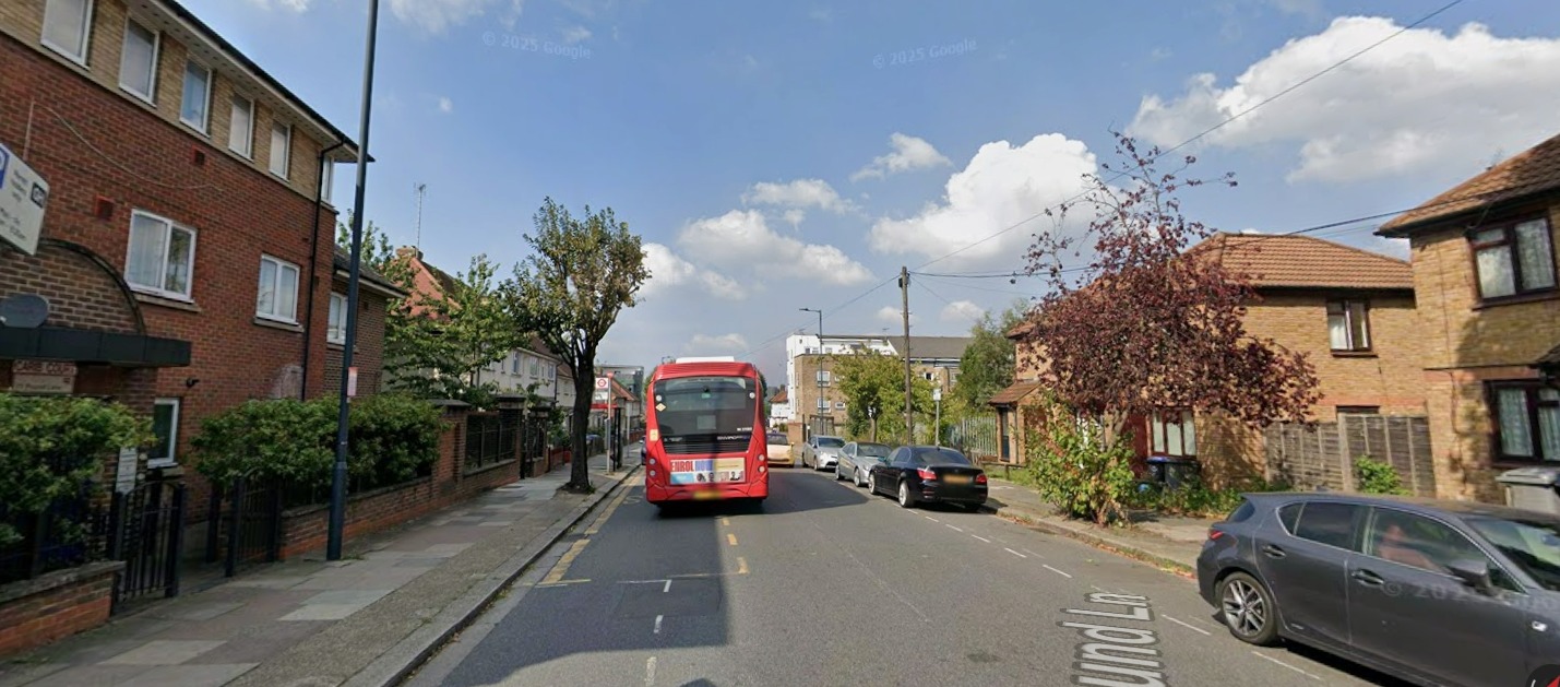 A red double-decker bus on a street lined with houses and parked cars.