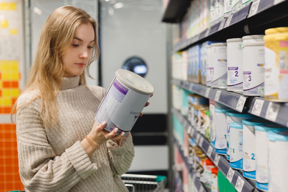 A young woman in a beige sweater looks closely at a can of infant formula in a supermarket aisle.