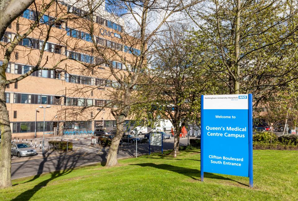 The Queen's Medical Centre Nottingham, a large brick hospital building, with a blue sign in the foreground welcoming visitors to the campus via the Clifton Boulevard South Entrance.