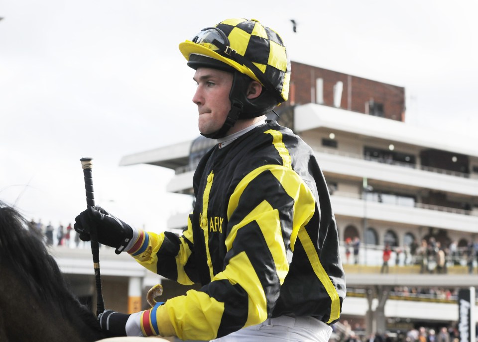 Amateur jockey Mr. Sam Lee in his racing silks, wearing a black and yellow checkered helmet and jacket, holding a riding crop.