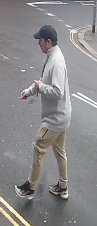 A person in a gray sweatshirt, tan pants, and black baseball cap walks across a paved surface.