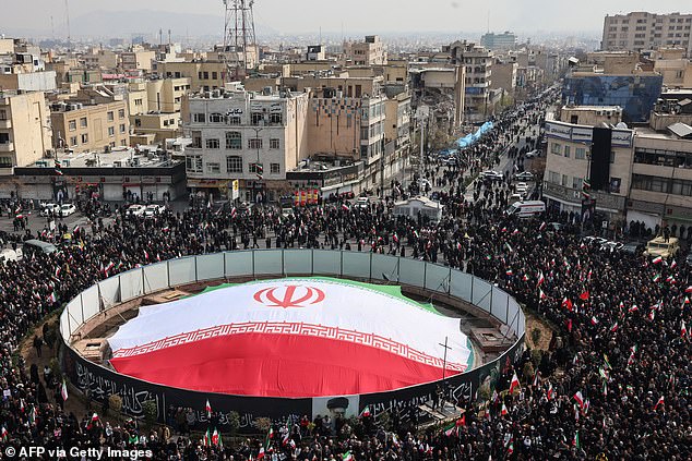Iranians attend the funerals of Iran's Revolutionary Guards Corps (IRGC) commanders, army commanders and others killed in the early days of the United States and Israeli strikes on Iran, with a damaged building visible in the background, at Enghelab Square in Tehran on March 11
