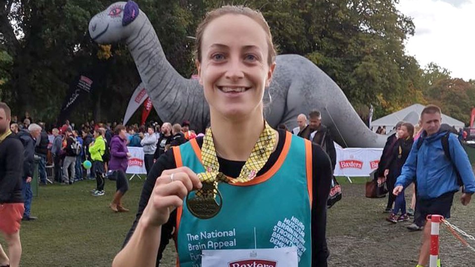 Woman in a teal bib with "The National Brain Appeal" and "Baxter's" printed on it holds up a medal.