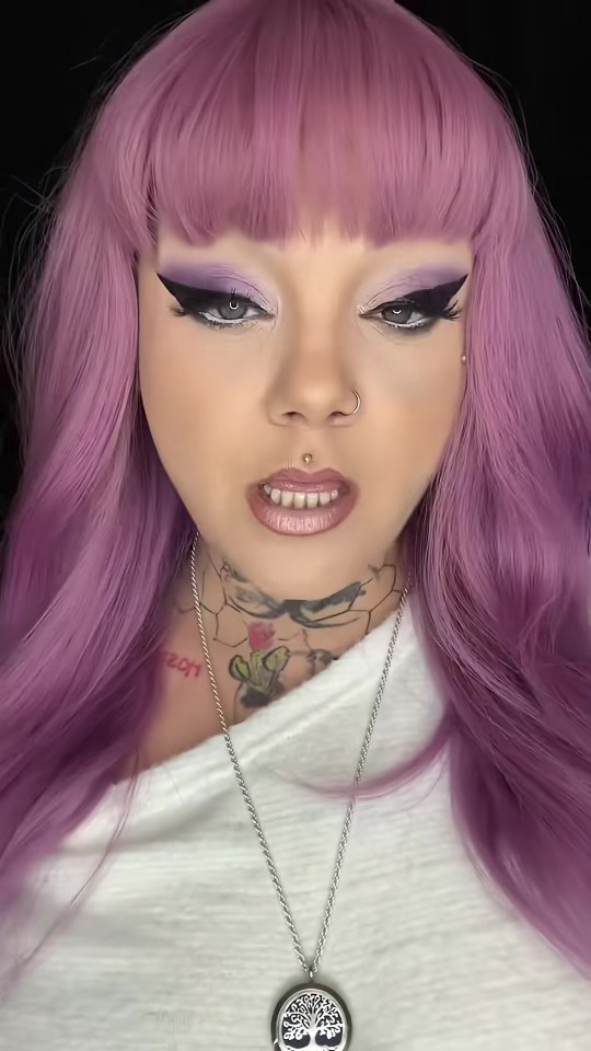 A woman with purple hair, purple eyeshadow, black eyeliner, a nose ring, and a philtrum piercing. She wears a white top with a tree of life necklace and tattoos on her neck.