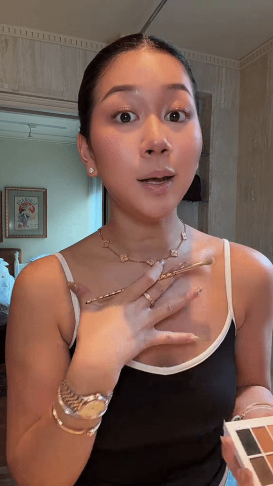 A woman in a black and white tank top holds an eyeshadow palette and a makeup brush.