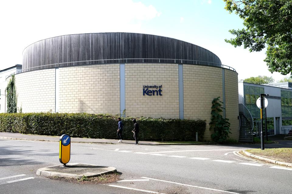 University of Kent building with brick and wood facade, green hedges, and two people walking by.