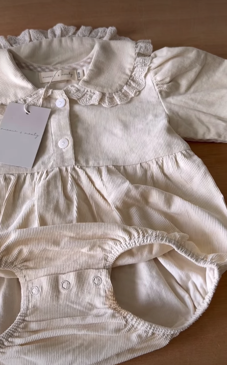 A light beige baby romper with a ruffled collar, long sleeves, and snaps at the crotch.
