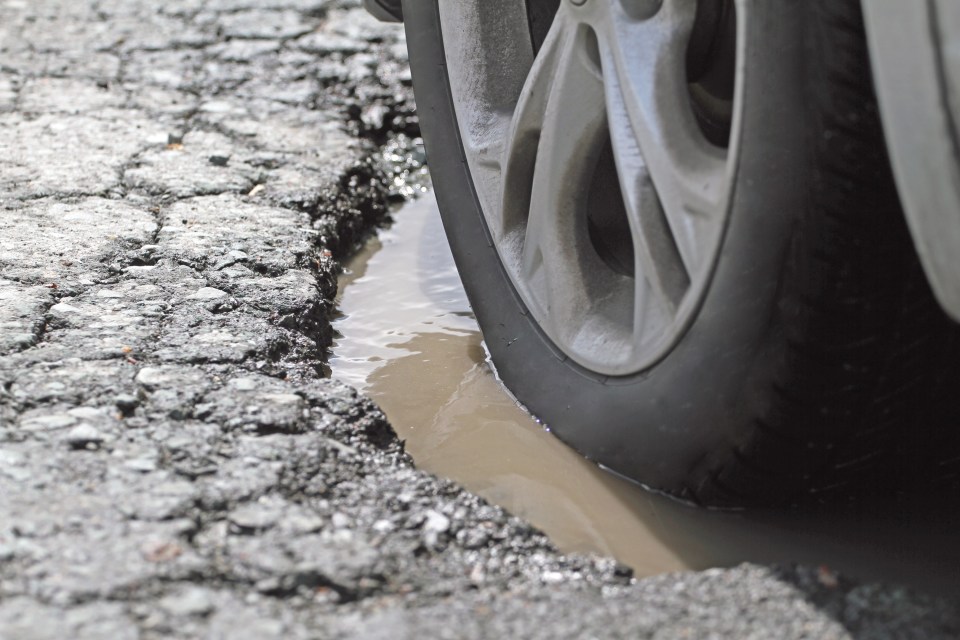 A car tire and alloy wheel are slowly driving through a pothole filled with murky water on cracked asphalt.