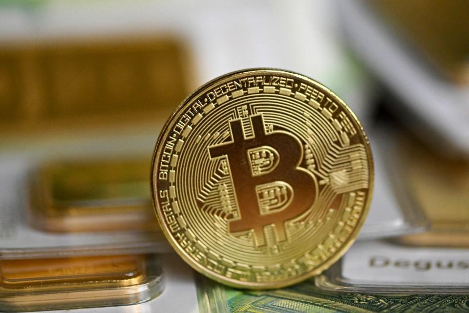 A gold Bitcoin coin standing on 100 euro notes, with two gold bullion bars in the background.