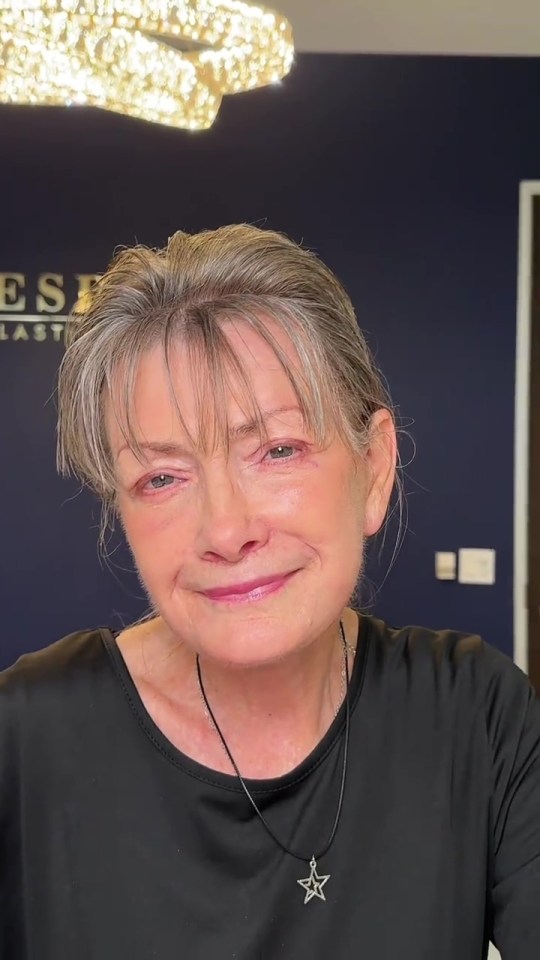 A woman with short grey hair and bangs smiles, wearing a black shirt and a silver star necklace.