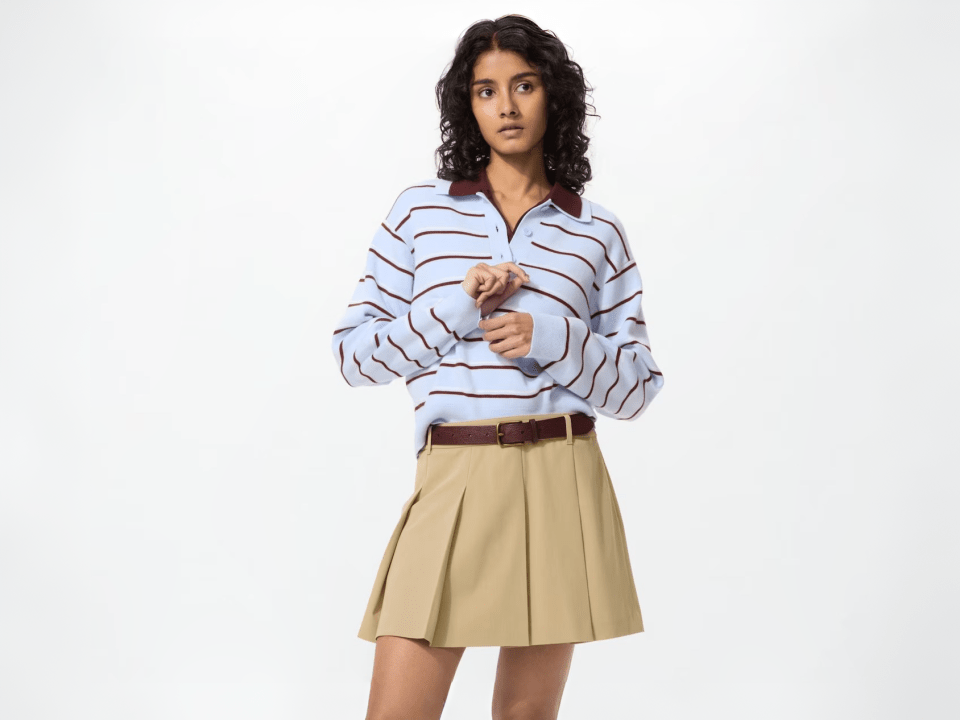 A woman wears a light blue striped long-sleeved polo shirt and a pleated tan skort with a brown belt.
