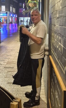A man standing outside at night, holding a dark coat, with a blurry street and buildings in the background.
