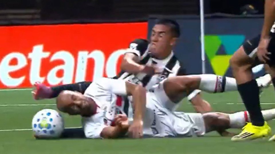 Two soccer players on the ground after a tackle, one in a white jersey and the other in a black and white striped jersey, with a soccer ball nearby.
