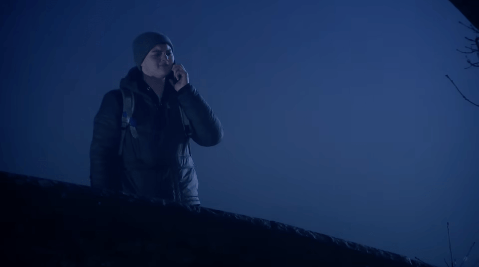 A man in a hat and winter jacket talks on a phone against a dark blue background.