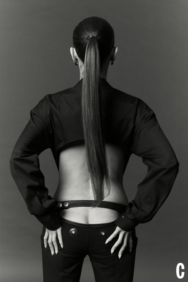 Maura Higgins from The Traitors US, seen from behind, wearing a cropped black shirt and black pants that expose her lower back, with her hair in a long ponytail.