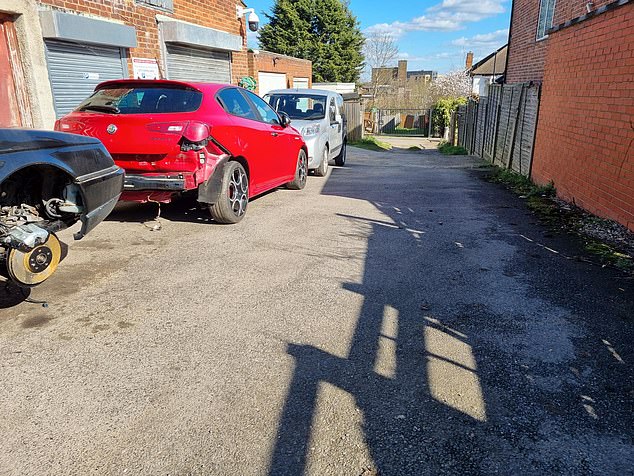 The lane with cars along one side outside the garages, including one with no rear bumper and one with its wheel removed