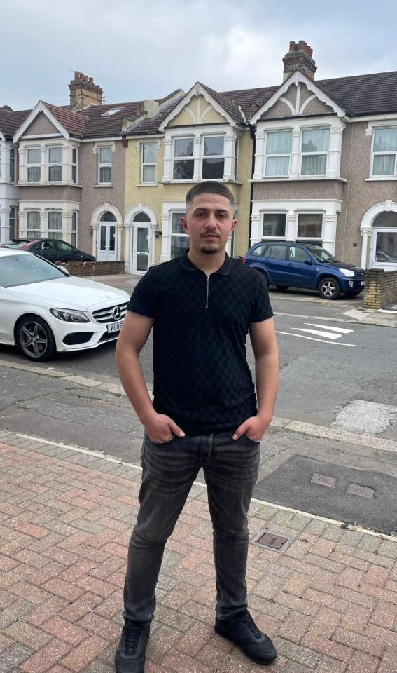 Renols Lleshi, a 19-year-old labourer, stands on a paved sidewalk with residential houses and parked cars in the background.