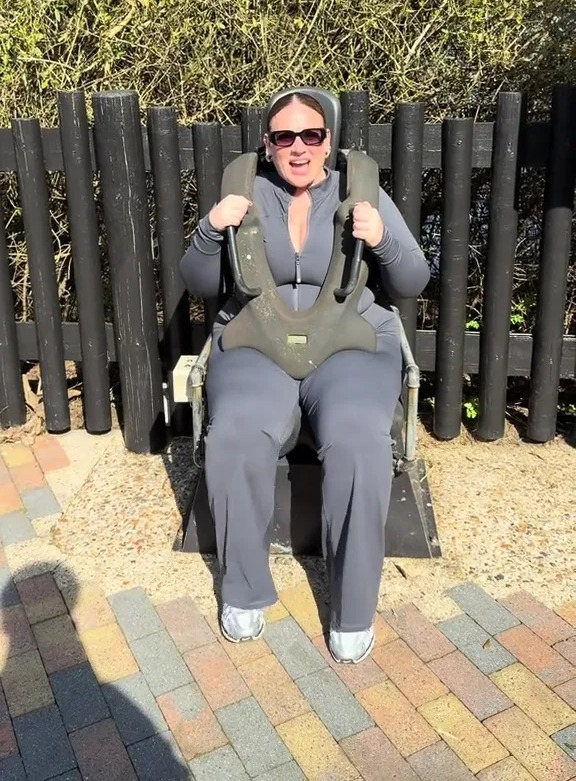 A woman in a gray tracksuit smiles, seated in a roller coaster seat with a safety harness.