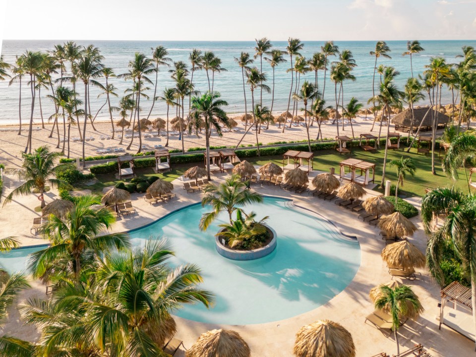 An aerial view of a resort with a large swimming pool, palm trees, and a sandy beach leading to the ocean.