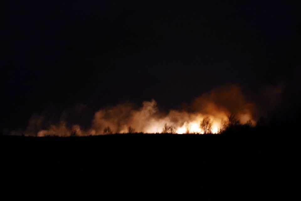 A moorland fire glowing at night, silhouetting trees and emitting smoke.